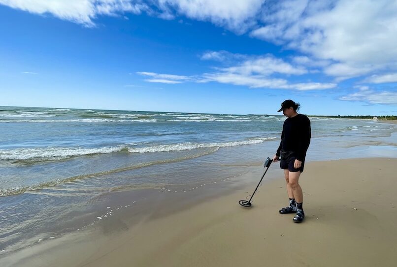 Christopher on a sunny beach, holding a metal detector, looking for treasure.